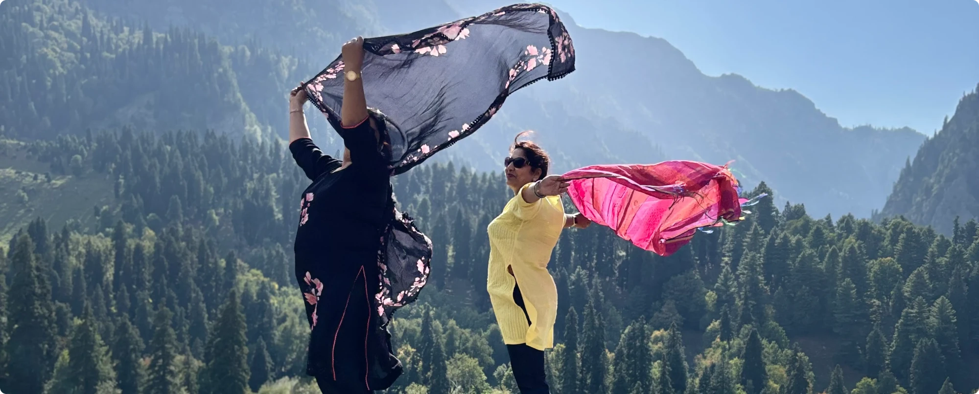 Two Sahelis letting their dupattas fly in the wind above a pine valley